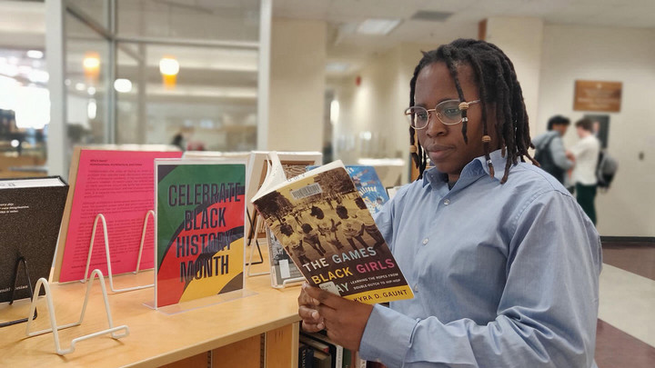 Moyo has dreadlocks with beaded hair accessories and wears gold framed glasses and a blue dress shirt. She is reading The Games Black Girls Play from the Celebrate Black History Month book display at the Mearns Centre for Learning – McPherson Library.