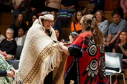 Indigenous woman wearing a woven blanket accepts a beaded feather gift from another woman wearing a red and black Indigenous form line shawl.