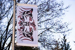A white flag with red and black artwork is installed on a black flag pole along a walking trail on campus.