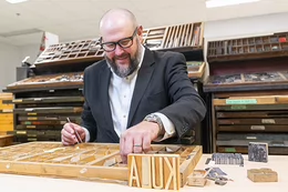 Matt Huculak is wearing a dark suit jacket, sitting at a table, with his hands in a wooden tray of metal typeface with “KULA” in large wooden blocks standing upright in front of the tray.