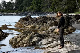 A student standing on a rocky beach surrounded by trees.