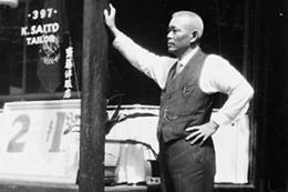 An older man in a vest and tie leans against a storefront. Powell Street, Vancouver, c. 1920.