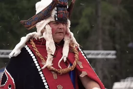 Hereditary Chief Frank Brown of the Heiltsuk Nation sits in a canoe, wearing traditional regalia that includes a red and black button blanket and cedar headdress. Behind him, there is ocean, sky, and green trees.