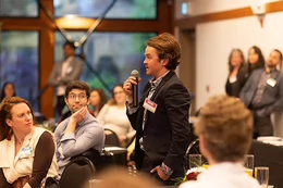 A standing man holds a microphone, asking a question, in a room full of people.
