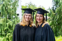 Two students stand together on campus wearing black convocation caps and robes.