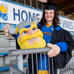 A student wearing regalia and a mascot outfit at an outdoor field.