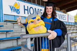 A student in a mascot outfit and grad regalia in the stands of an outdoor field.