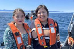 Two young Inuit women in lifejackets stand on a boat with the sea in the background.