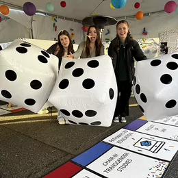 Three students pose with three life size dice on a UVic themed monopoly board, under a white event tent decorated with colourful round lanterns.