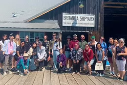 A large group of field school students poses for a group photo in front of a wooden structure featuring a sign that says Britannia Shipyard.