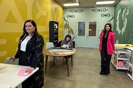 Three women stand and sit in an art lab. The walls are painted yellow and green with Indigenous artwork.