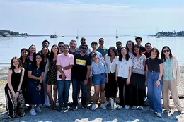 A group of students stand on the beach at Cadboro Bay, BC smiling with a view of a calm ocean and boats in the background.
