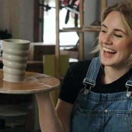 Smiling, blonde woman in overalls holds up top of pottery wheel with recently made cup on it.