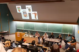 A large class sits in a lecture hall as six people sit at the front. Indigenous artwork is displayed on the projection screen behind them.
