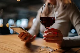 A person holds a cell phone and a glass of red wine in the other hand.