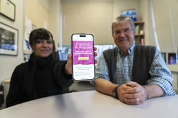 Two people sit at a table as one holds up a phone screen showing a new website.