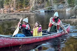 A student and two children paddle a canoe on a calm lake.