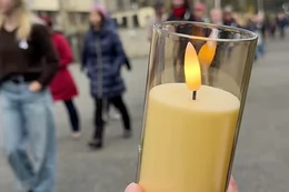 A yellow battery operated candle glows in a yellow glass holder. A long line of people are seen walking along a pathway on campus in the background.