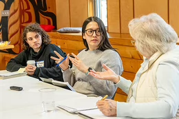 Three women of different ages sit at a table, gesturing with their hands as they speak.