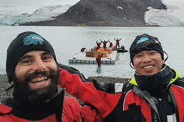 Two men wearing toques and red jackets pose together on a rocky shore. Behind them a group of people stand in the water and on a dock.