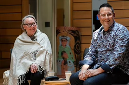 Siila Watt-Cloutier and Dr. Ian Mauro sit side by side in chairs at the ceremonial hall in First Peoples House at UVic. Both are smiling and looking to the right, engaged with someone in the audience. Watt-Cloutier is wrapped in a ceremonial blanket, and Mauro wears a floral shirt. Both wear glasses. A small table with three cups of water sits between them.