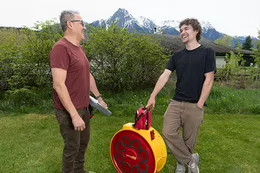 Energy Advisor Richard Joseph and mentee Ari Orrantia stand beside a blower door fan, smiling and talking outdoors with snow-capped mountains in the background.