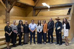 Ten people stand in a line side by side in the Stz'uminus Big House, with wooden bench seats behind them.