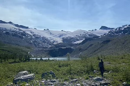 A student with backpacking gear hiking beside a stream toward an alpine glacier in the background.