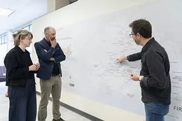Three people look at a large map on a white wall.