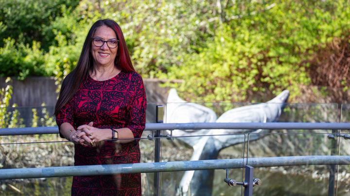 Associate Vice-President Indigenous Robina Thomas stands on the walkway over a pond behind First Peoples House. The Whales Tail sculpture is visible behind her. She is wearing a red dress.
