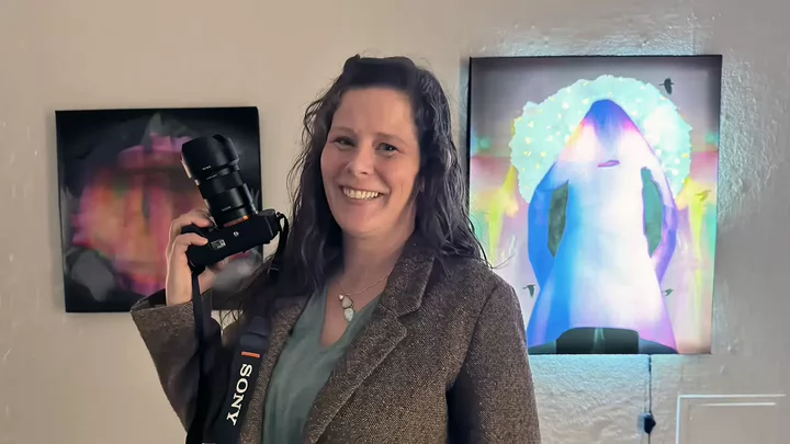Beth holds a digital SLR camera smiling, behind her are two pieces of art on the wall