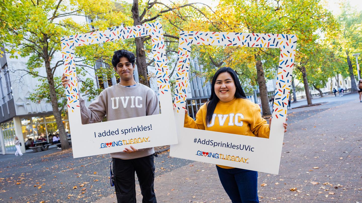 A male and female student are standing smiling and posing in frames decorated in sprinkles. The male student on the left holds a frame that says I added sprinkles! The female student on the right's frame says #AddSprinklesUVic Giving Tuesday