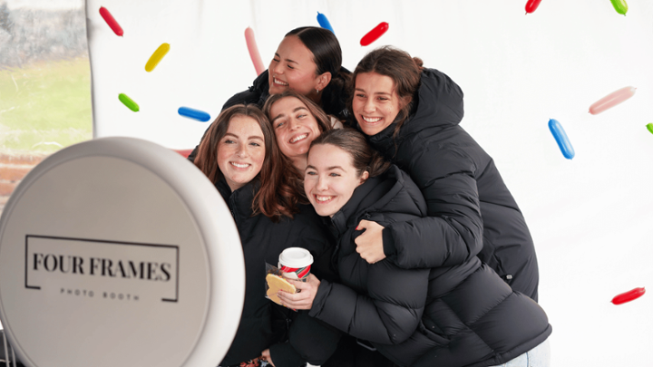 A group of students smile at the camera in a photo booth, a colored sprinkle background is behind them