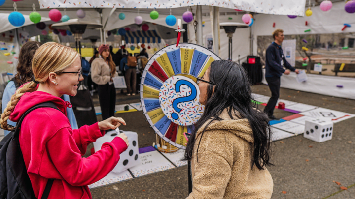 two students are smiling talking to each other in front of a colorful wheel, the background behind them is a tent with colorful decorations and a life-size game board with giant dice and people playing games