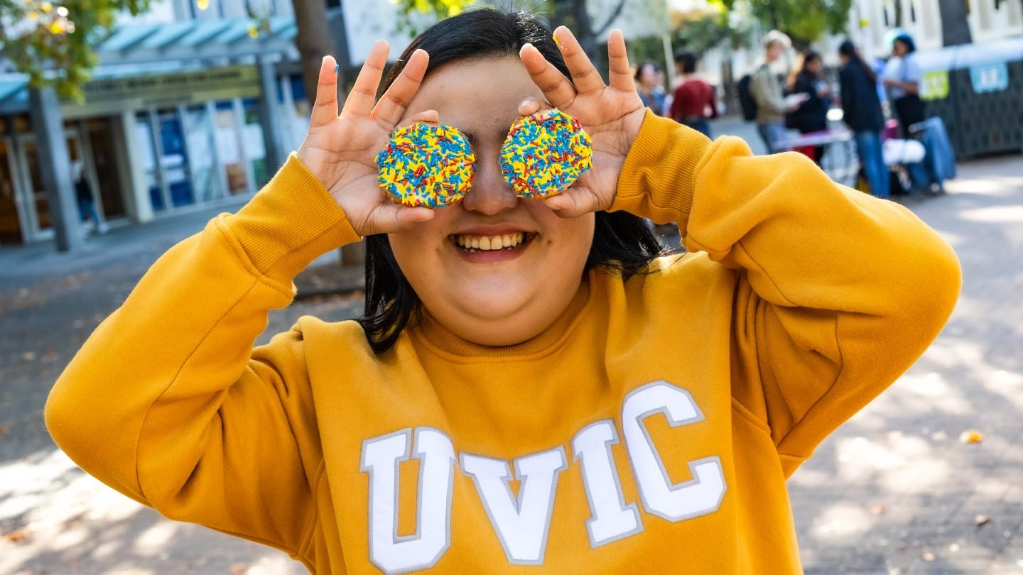 Student in bright yellow UVIC sweater holding two sprinkle cookies up to her eyes