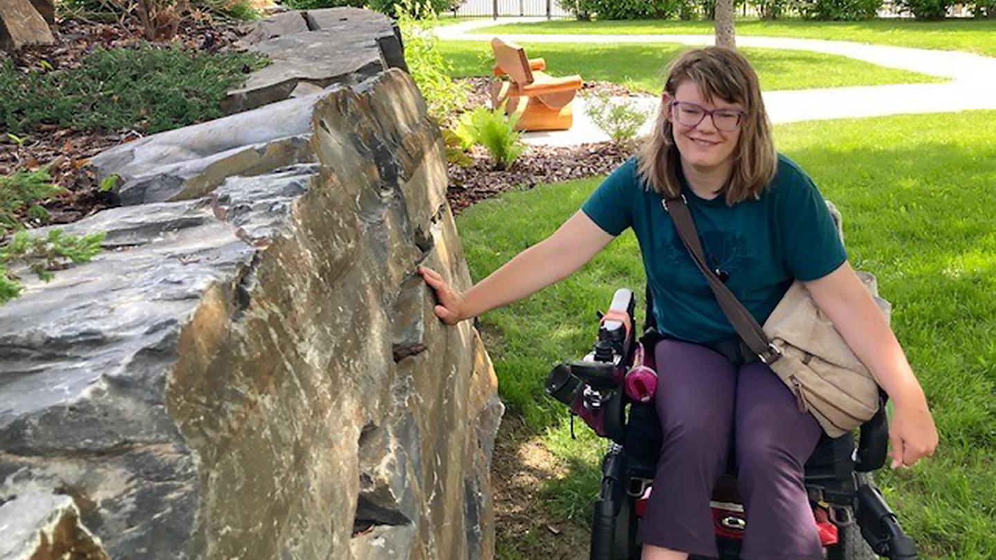 A woman in a wheelchair on a lawn smiles at the camera while touching a rock wall to her left