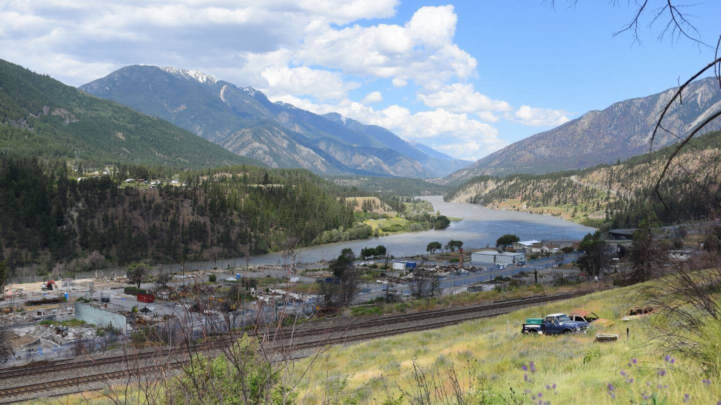 A rail line and burned remains of buildings in Lytton, BC in the foreground, with a river valley and mountains in the background.