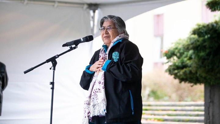 Elder May Sam standing in front of a microphone providing a land blessing.