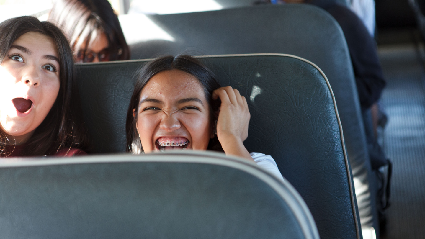 Two young women are making silly faces and laughing, while sitting on a grey bus seat. There are other people around them not in focus. 