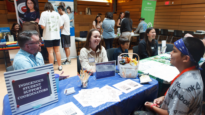 Students meet with UVic staff and faculty in the Ceremonial Hall, sitting at tables, a sign reads "Indigenous Student Support Coordinators" beside participant.