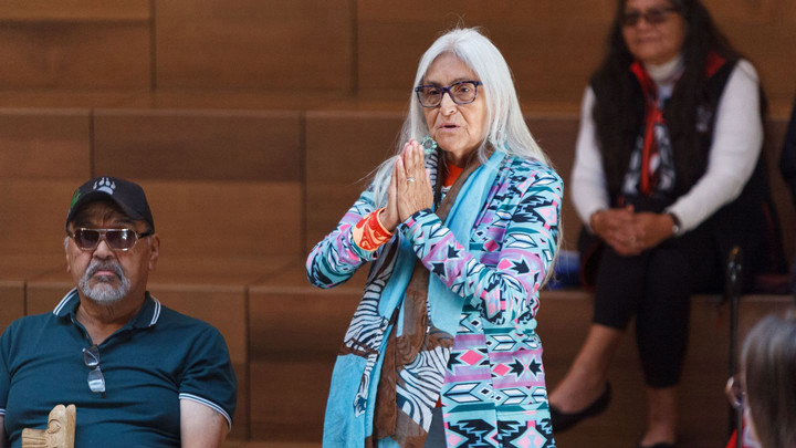 Elder Kathy Horne standing in First Peoples House, her hands together in prayer. Beside her sits Elder Doug LaFortune.