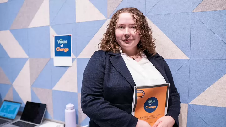 Nadiya in a business suit holding a document. On wall behind is a poster for Voices of Change.