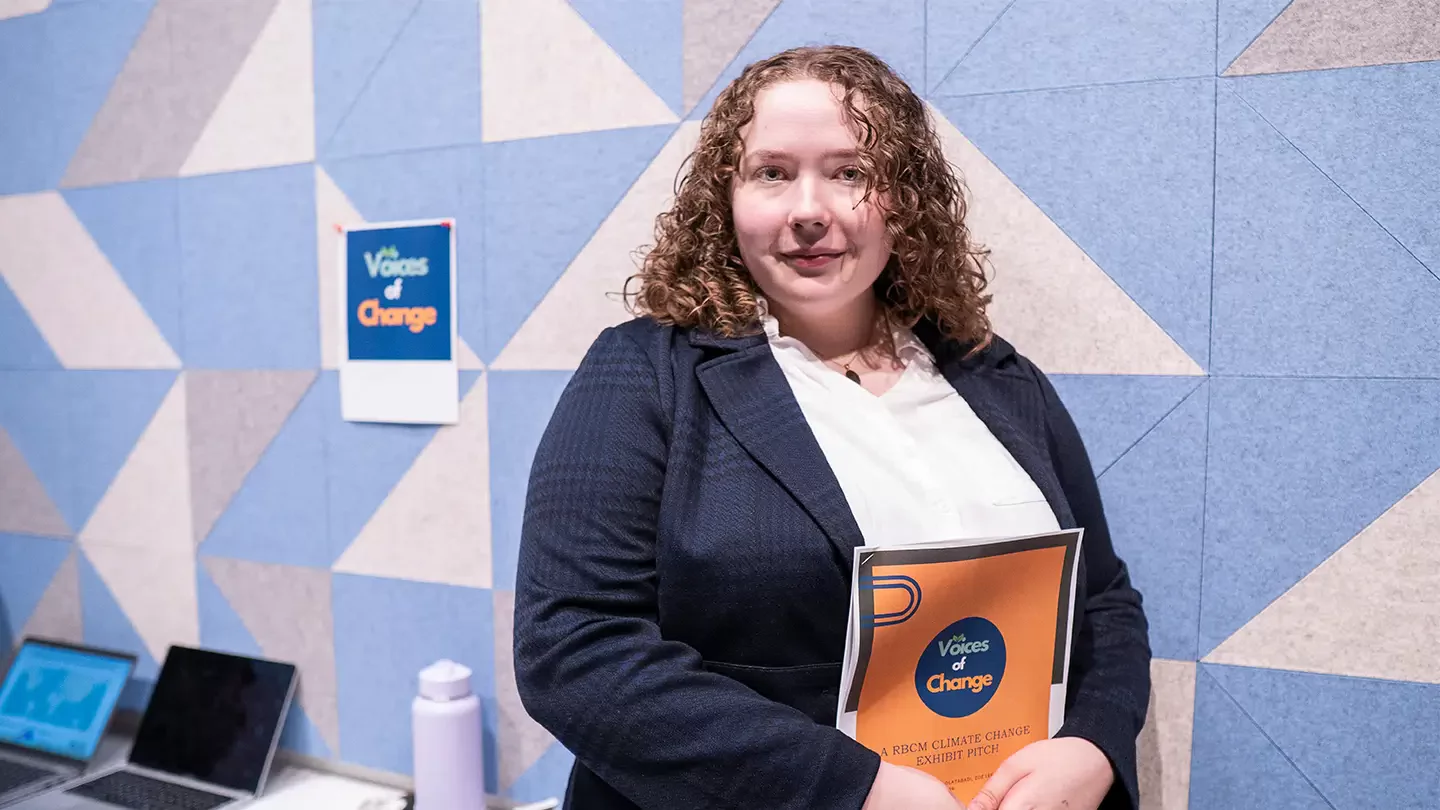 Nadiya in a business suit holding a document. On wall behind is a poster for Voices of Change.