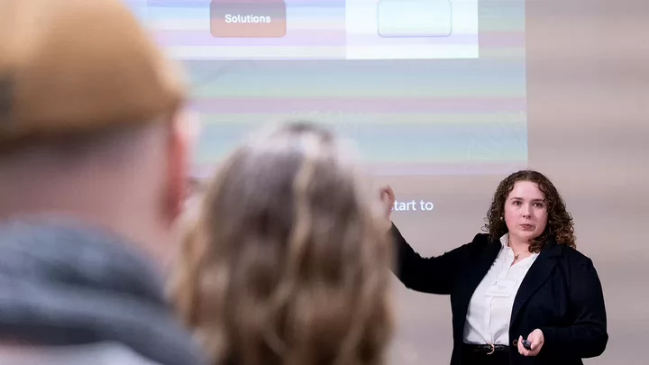 A person in a business suit pointing to a presentation screen, with audience members' heads visible in foreground
