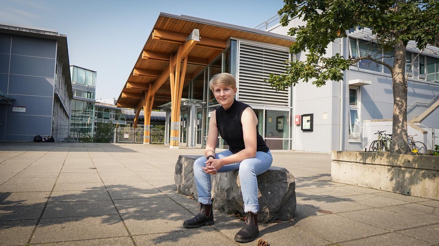 Student with cropped blonde hair and blue eyes sitting on stone bench in front of university building