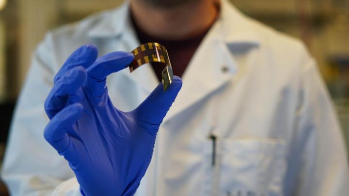 An out of focus student is holding an in focused flexible solar cell with blue gloves on.