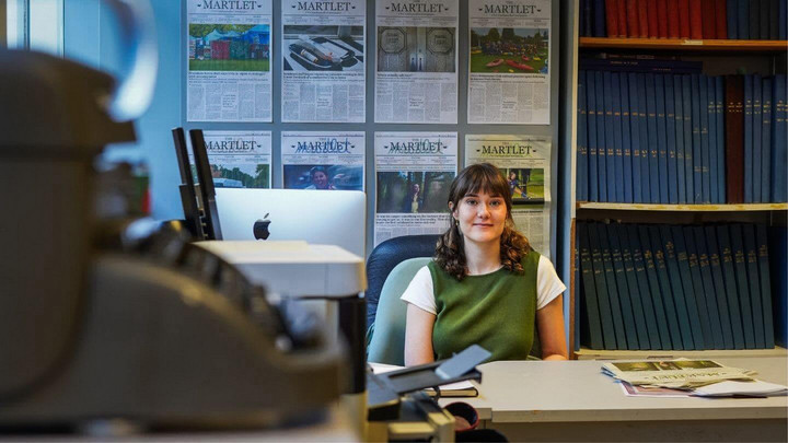 A person sitting at a desk with front pages of the Martlet newspaper displayed behind them and an old typewriter in the foreground