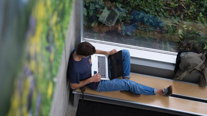 A student sits on a bench next to a window working on their computer.