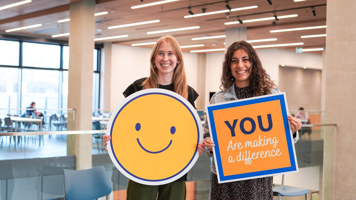 Two female students stand smiling. One is holding a large yellow smiley face and the other is holding a sign that says "you are making a difference"