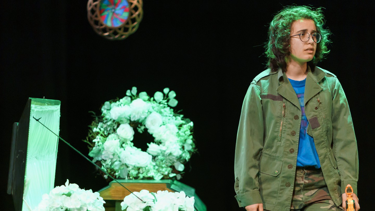 Zoe on stage in the play The Wake wearing army fatigues standing next to an open coffin